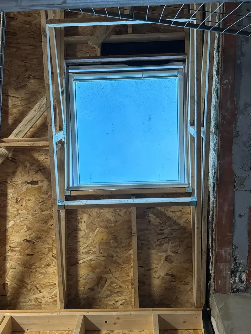 Fenêtre de toit vue depuis les combles, encadrée par un chevêtre en bois neuf, entre des panneaux OSB